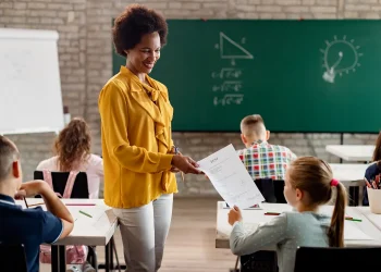 Professora entrega atividade para aluna em sala de aula durante avaliação, representando os desafios e a realidade da profissão docente.