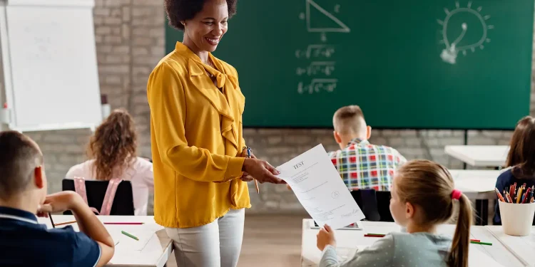 Professora entrega atividade para aluna em sala de aula durante avaliação, representando os desafios e a realidade da profissão docente.