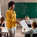 Professora entrega atividade para aluna em sala de aula durante avaliação, representando os desafios e a realidade da profissão docente.