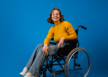 Jovem sorridente em cadeira de rodas com blusa amarela em fundo azul.