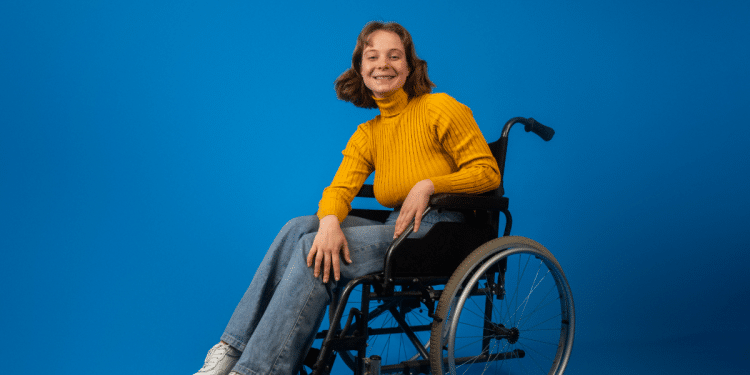 Jovem sorridente em cadeira de rodas com blusa amarela em fundo azul.