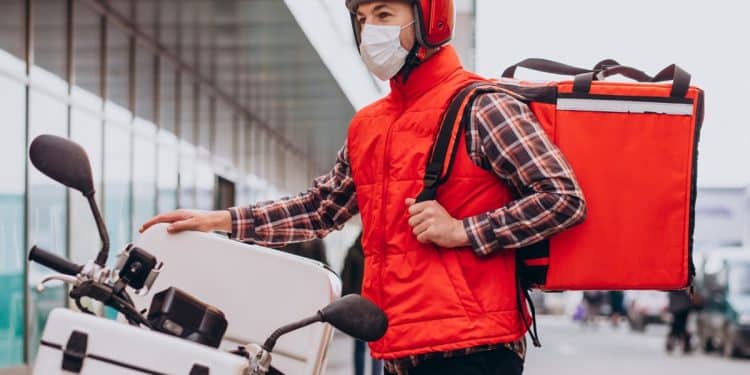 Entregador de capacete e colete vermelhos ajusta a mochila térmica ao lado de uma moto na rua