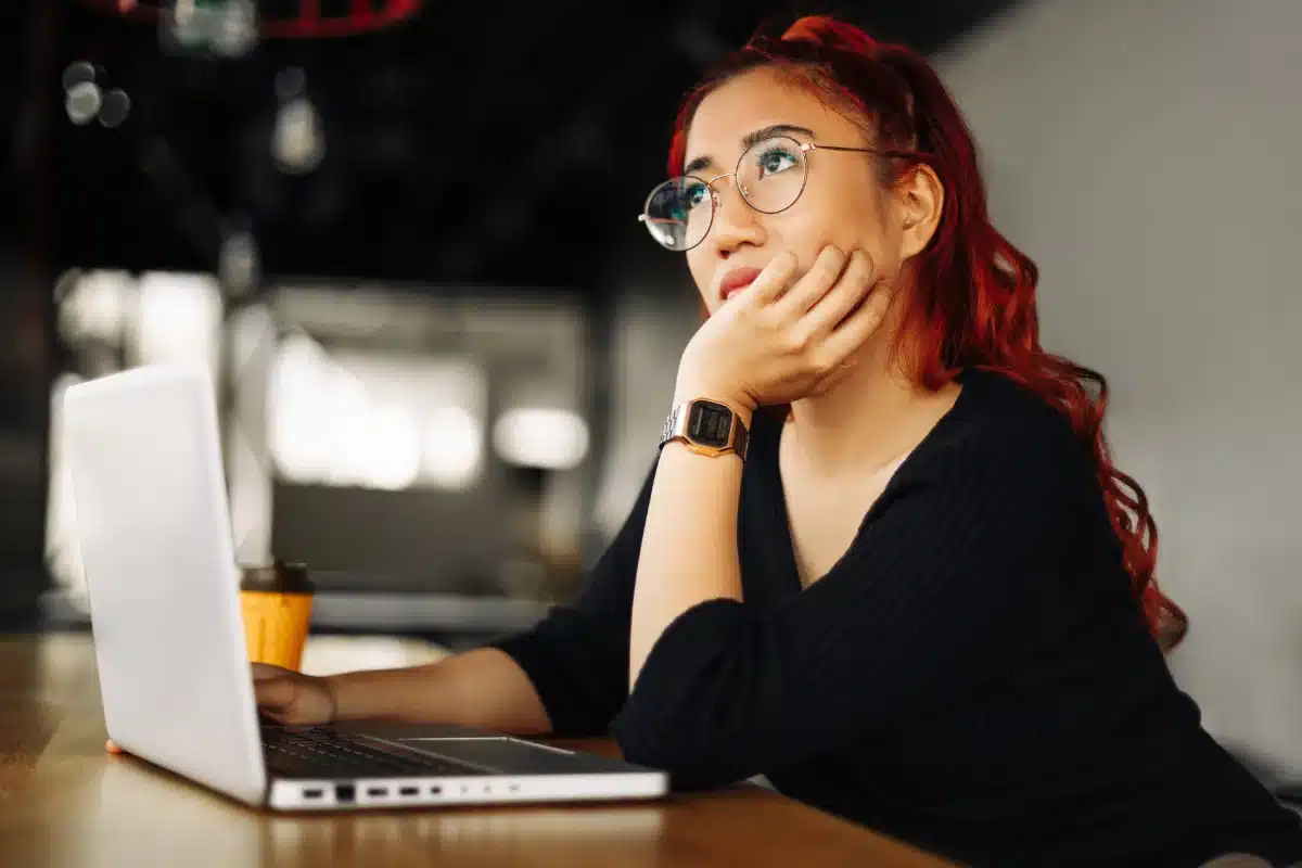 Mulher em dúvida Mulher com cabelo vermelho pensativa, apoiando o rosto enquanto olha para o computador