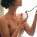 Jovem mulher segurando perfume vintage perto do pescoço, usando brinco dourado circular e vestido bege, simbolizando a dúvida entre “flagrante” e “fragrante”.