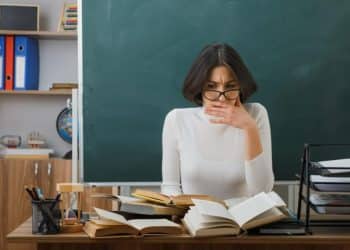 Mulher de óculos com a mão na boca, em expressão de surpresa, diante de livros abertos sobre a mesa