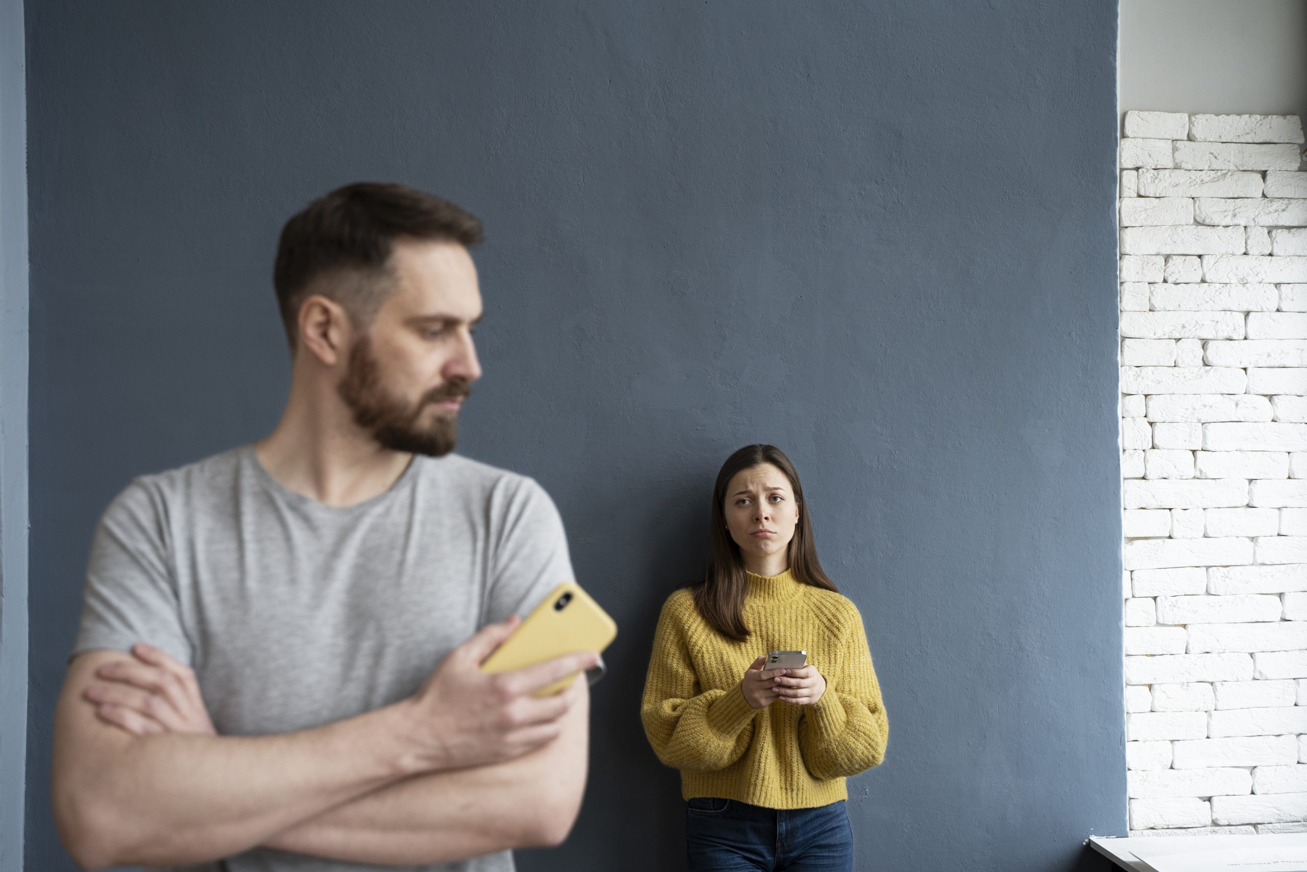 Psicologia e narcisismo. Homem de braços cruzados segurando um celular amarelo e olhando para o lado, enquanto uma mulher ao fundo, usando um suéter amarelo, segura um celular e aparenta estar triste ou preocupada