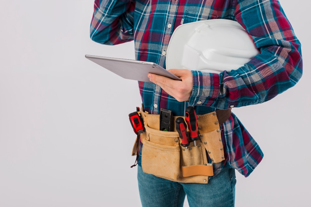 Trabalhador técnico ou engenheiro segurando um tablet e um capacete branco, vestindo camisa xadrez e cinto de ferramentas com chaves de fenda e fita métrica