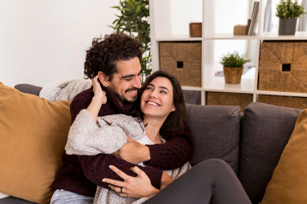 Jovem casal feliz sentado na sala de casa abraçados e sorrindo