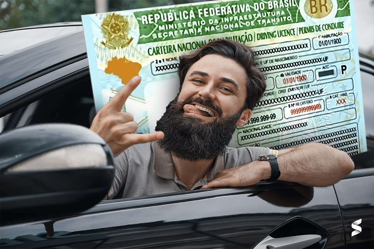 Homem sorridente dentro de um carro, fazendo um gesto de 'rock' com a mão e exibindo uma carteira de habilitação do Brasil em tamanho ampliado ao fundo