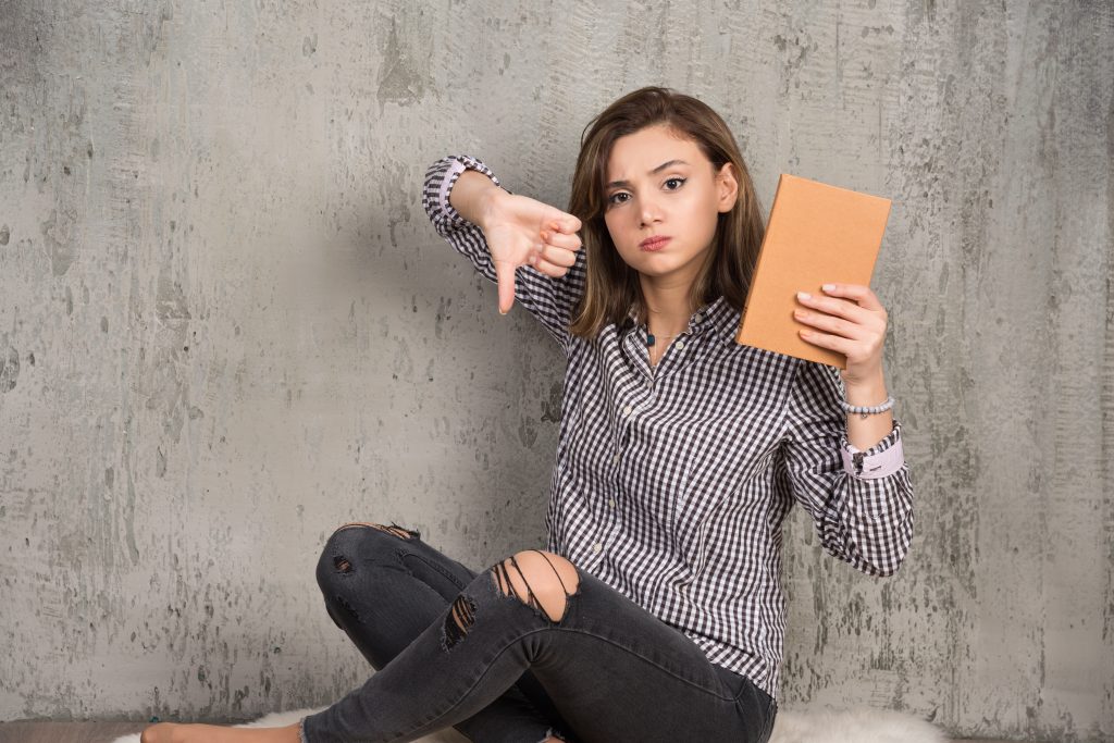 Female student with orange book giving thumbs down Hábitos que afastam a felicidade e, consequentemente, impedem o crescimento pessoal.