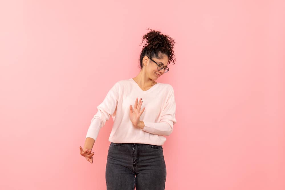 Mulher de cabelos cacheados e óculos, vestindo blusa clara e calça jeans, fazendo um gesto de recusa com as mãos, expressando desconforto, contra um fundo rosa
