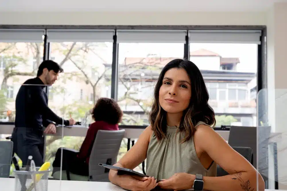 Mulher sorrindo em escritório moderno com ambiente colaborativo e profissionais ao fundo