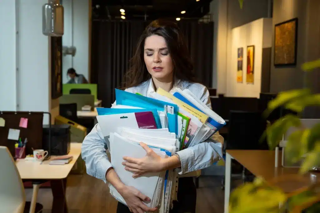 Sobrecarga de trabalho e seus impactos na vida pessoal e profissional. Mulher em escritório carregando pilhas de documentos, ilustrando o impacto da sobrecarga no trabalho e vida pessoal.
