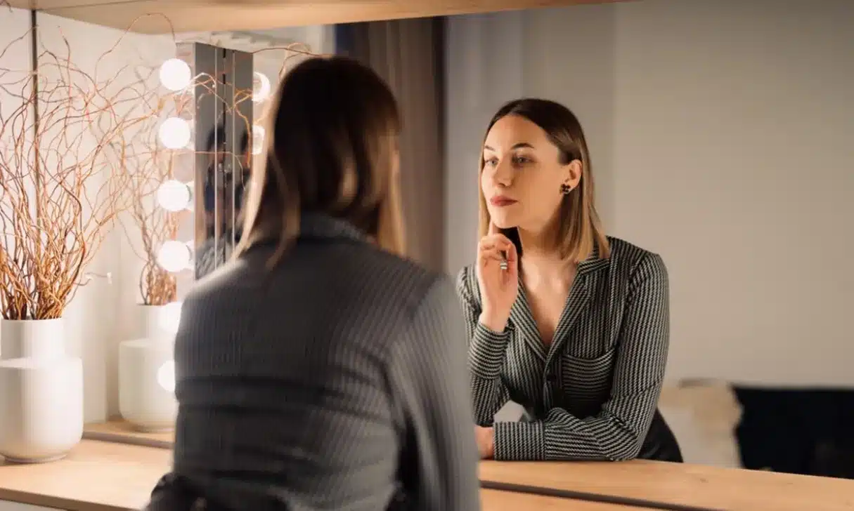 Como superar o desconforto inicial ao falar diante do espelho Mulher olhando para o espelho com expressão pensativa, representando a superação da resistência inicial na autoconversa