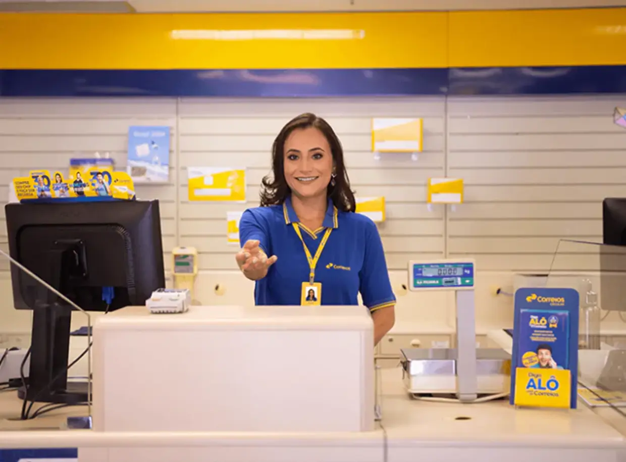 Atendente Comercial dos Correios em Atendimento Atendente comercial dos Correios atendendo no balcão de uma agência, com uniforme oficial e sorrindo para os clientes
