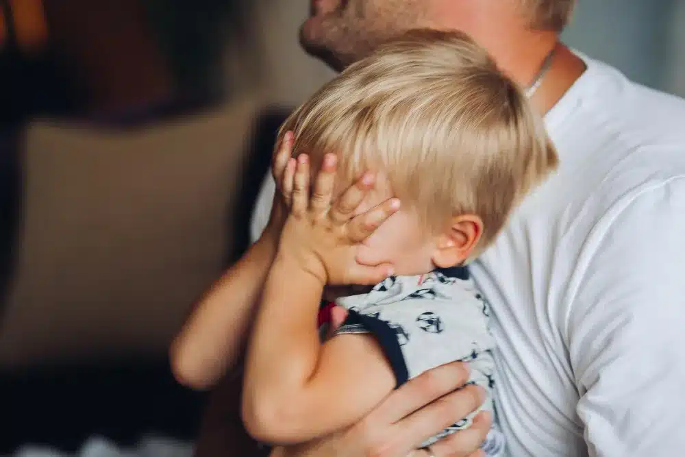 Criança cobrindo o rosto com as mãos enquanto é abraçada, simbolizando dificuldades emocionais na infância.