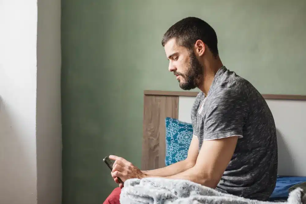 A dificuldade de pedir ajuda e suas raízes emocionais. Homem sentado na cama olhando para o celular com expressão reflexiva e preocupada.