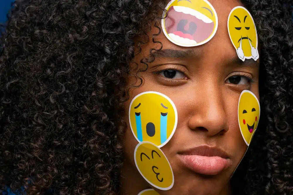 Mulher de expressão triste com adesivos de emojis variados no rosto, representando diferentes emoções.