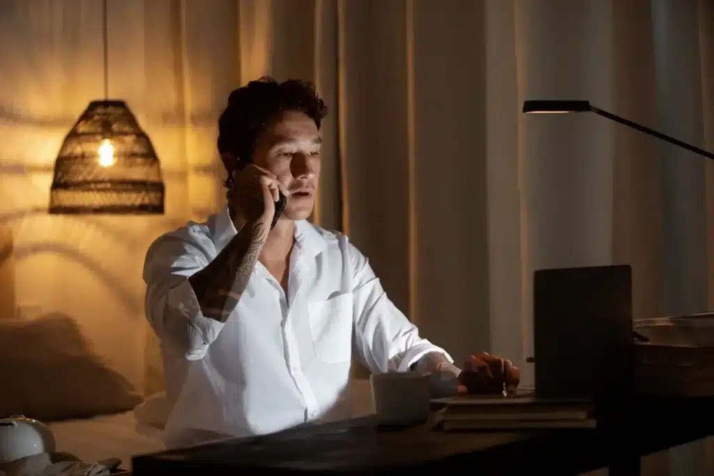 Hábitos Curiosos de Pessoas Muito Inteligentes Homem trabalhando à noite, falando ao telefone e usando laptop, representando hábitos curiosos de pessoas inteligentes