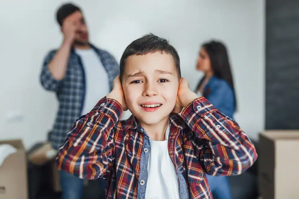 Criança cobrindo os ouvidos enquanto os pais discutem ao fundo, representando o impacto emocional do narcisismo parental.