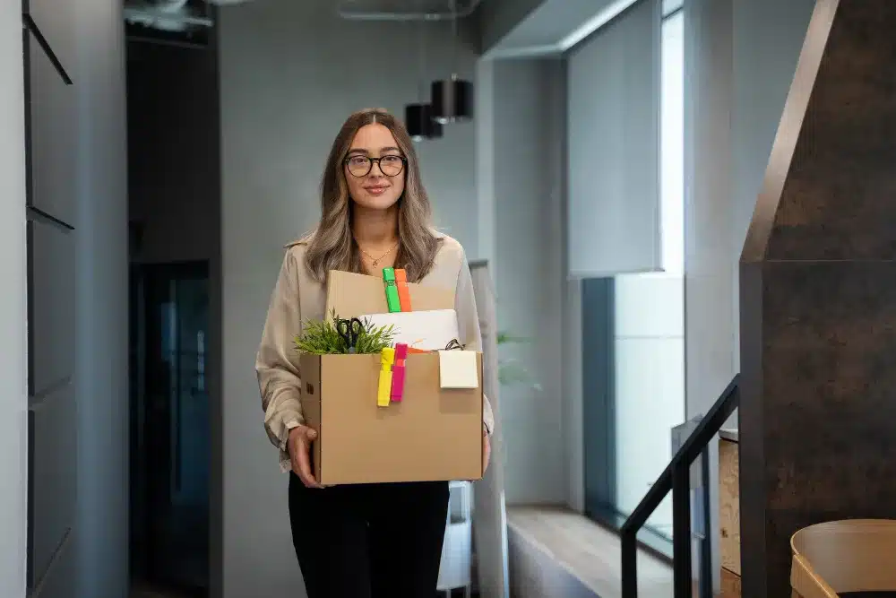Mulher saindo do trabalho com uma caixa de papelão, simbolizando a mudança de emprego.