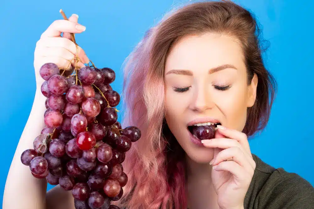 Uvas – Fonte Rápida de Energia para Treinos Mulher segurando um cacho de uvas e consumindo uma, destacando sua importância na nutrição esportiva.