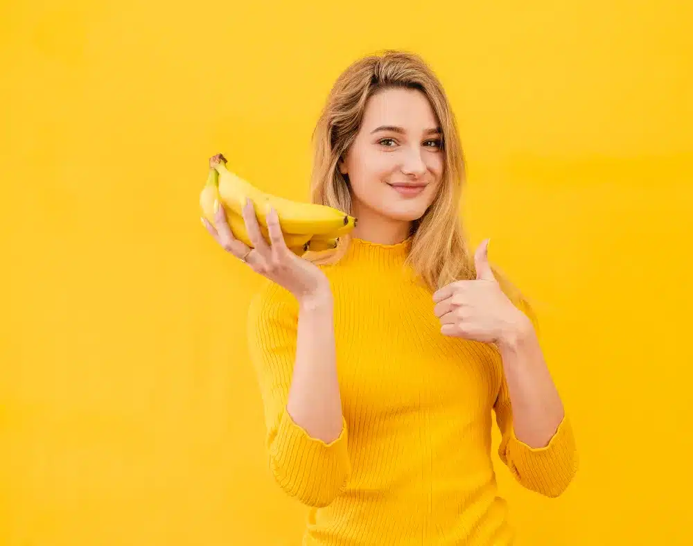 Mulher com Bananas – Fonte Natural de Energia Mulher sorrindo e segurando bananas, representando alimentação saudável e energia para treinos