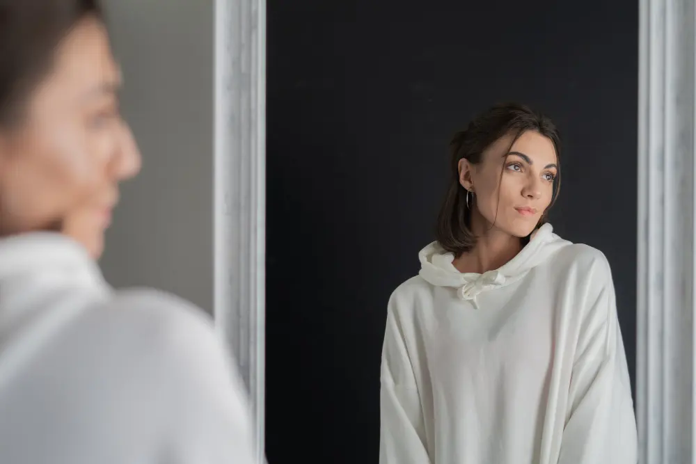 Reflexão narcisista: a busca pela validação Imagem de uma mulher olhando para o espelho, refletindo sobre si mesma. Ela parece pensativa, simbolizando a busca constante por validação e a necessidade de ser admirada.