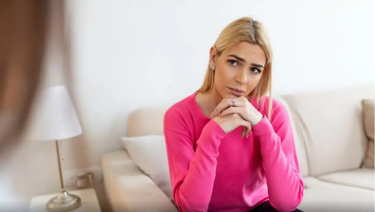 Os perigos da busca constante por aprovação Mulher sentada no sofá com expressão de preocupação, refletindo sobre a opinião dos outros e o impacto disso na sua vida