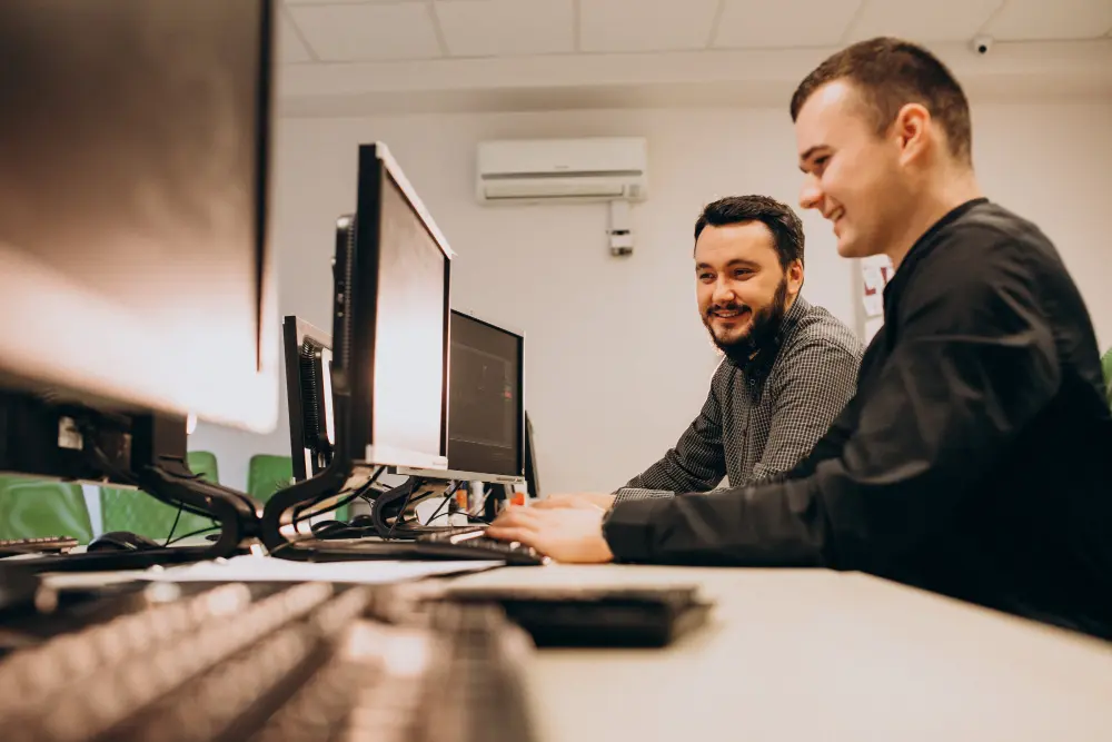 Dois profissionais em um ambiente de escritório, trabalhando juntos em frente a computadores, sorrindo enquanto colaboram