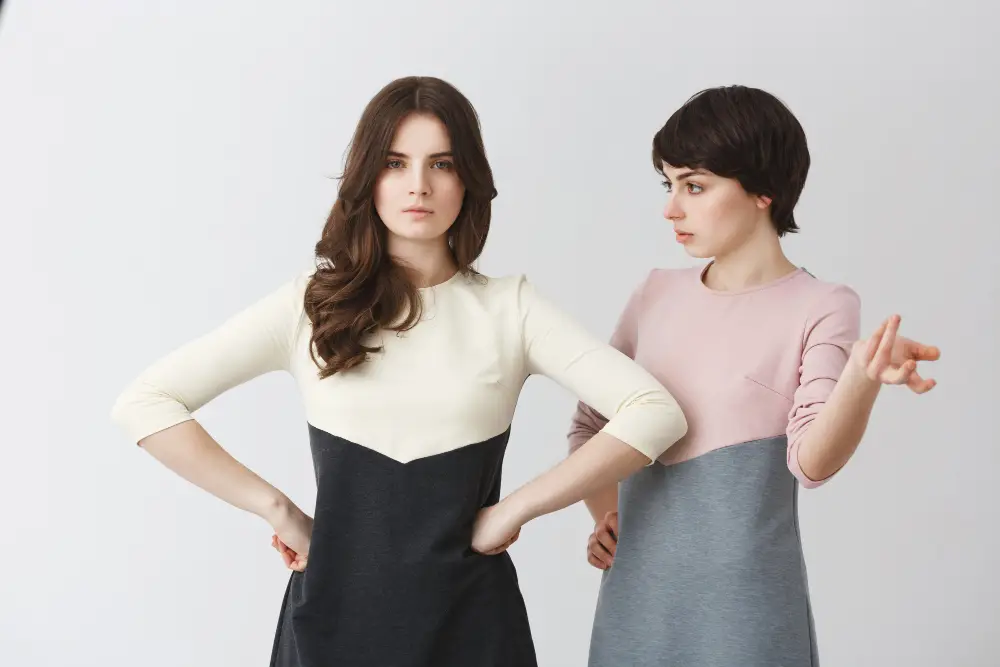 Duas mulheres em poses diferentes com vestidos bicolores, uma demonstrando uma expressão séria e a outra com uma postura gesticuladora, ilustrando um tema sobre atitudes egoístas