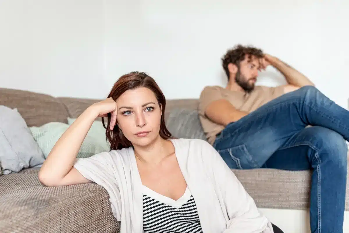 Casal sentado no sofá com expressões sérias, indicando possível distanciamento emocional.