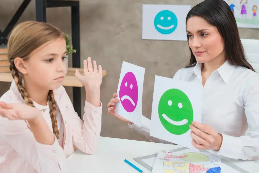 Como superar a dificuldade de expressar sentimentos? Psicóloga segurando cartões com expressões faciais enquanto criança demonstra indecisão sobre emoções.