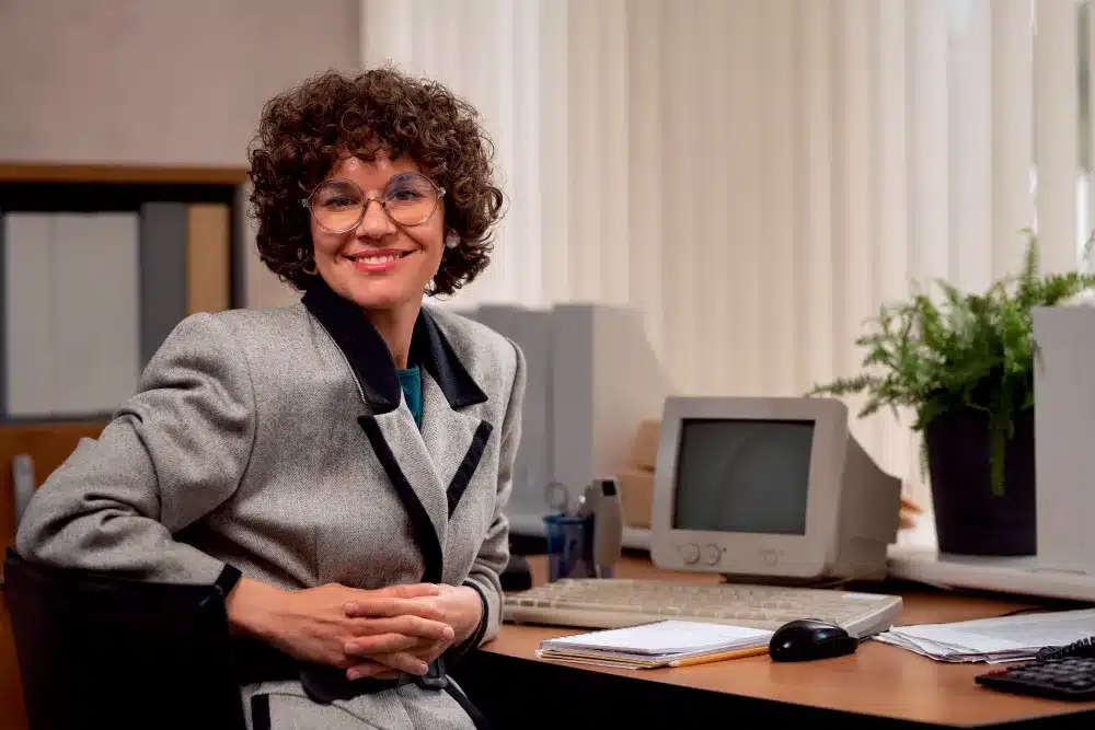 Técnico em Secretariado: profissional fundamental para a organização corporativa Profissional de secretariado em ambiente corporativo, sorrindo e com um computador à sua frente, representando a organização e eficiência no escritório