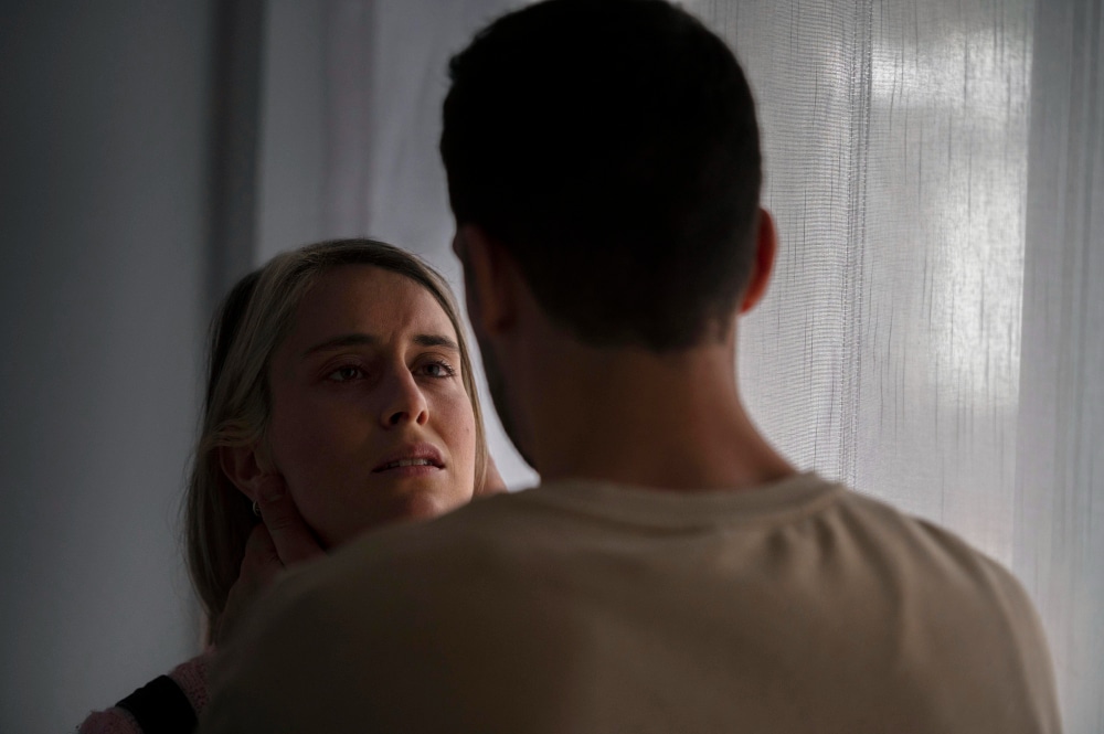 Mulher com expressão de angústia sendo segurada pelo pescoço por um homem, em um ambiente com luz baixa.