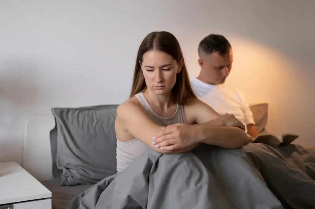 Impacto da manipulação emocional na autoestima Mulher com expressão preocupada, abraçando o próprio corpo, enquanto o parceiro está ao fundo, focado em um livro, refletindo uma possível tensão emocional no relacionamento.