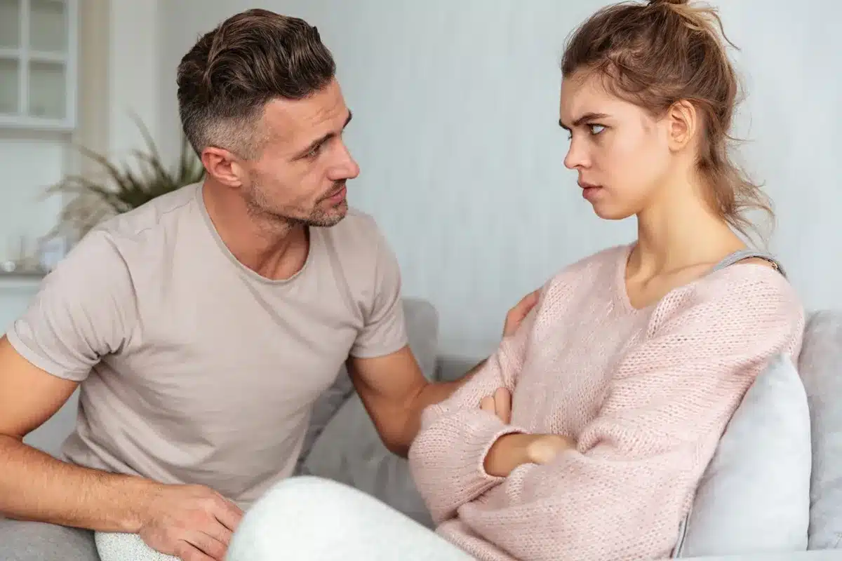 Casal em discussão, com um homem tentando se explicar enquanto a mulher se mostra desconectada emocionalmente.