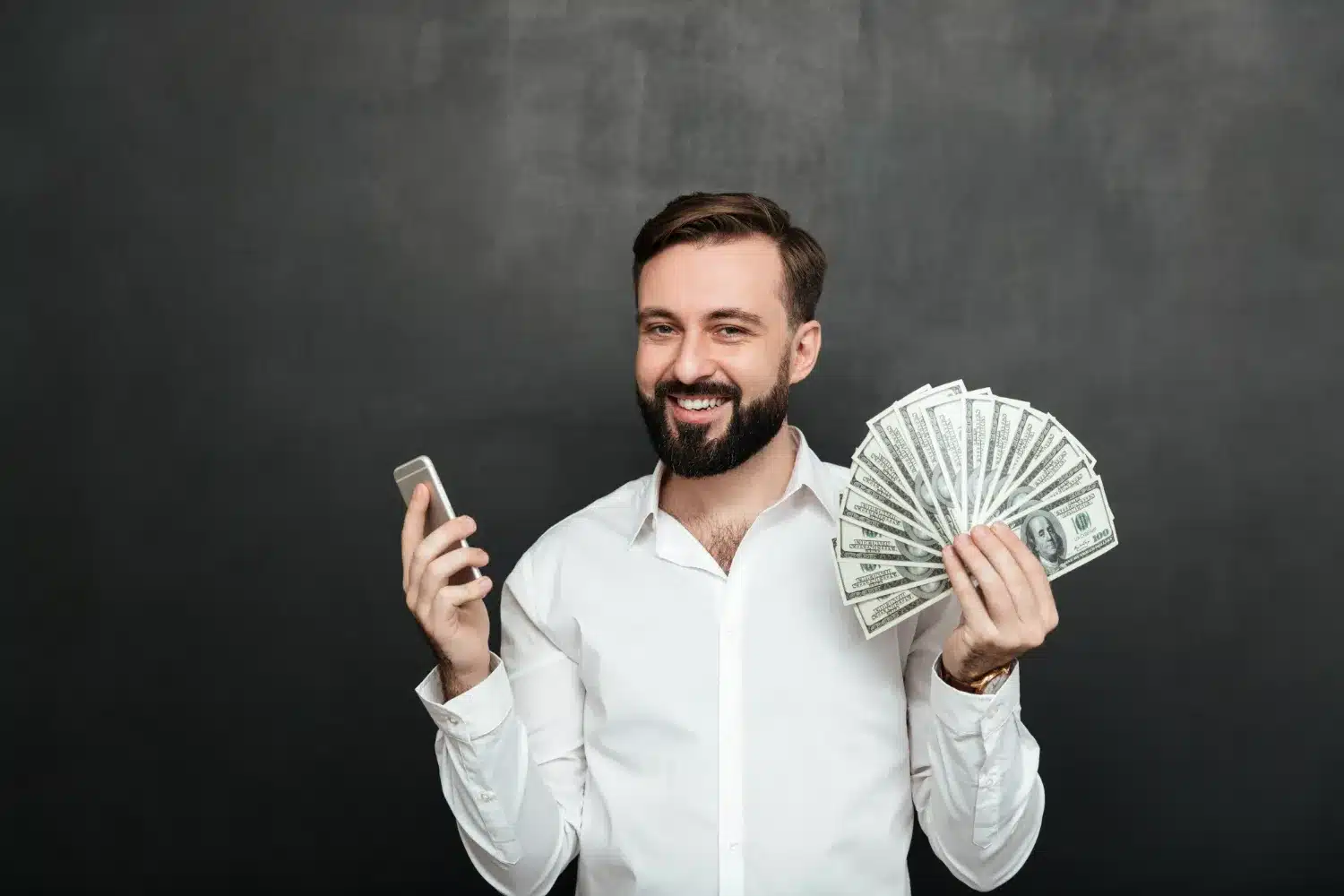 Homem sorrindo e segurando notas de dólar em uma mão e um celular na outra, simbolizando sucesso financeiro.