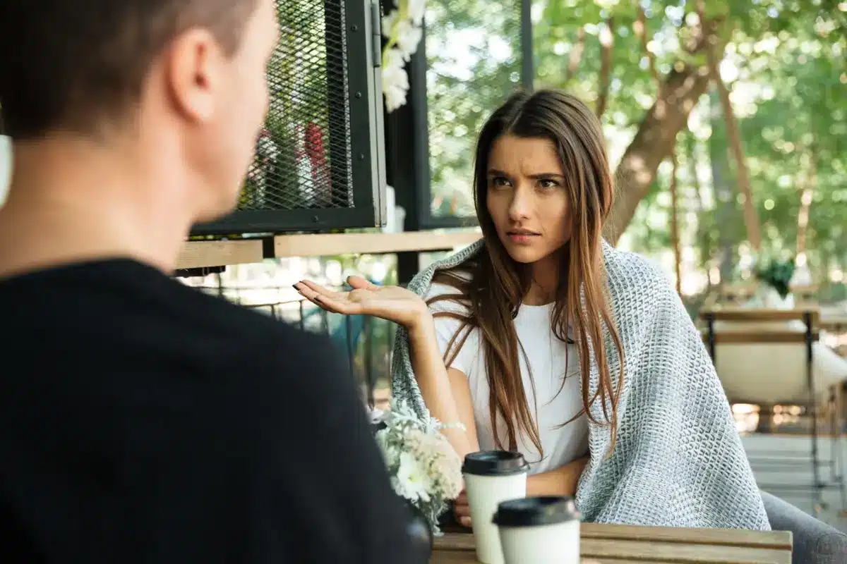 Mulher com expressão de desconfiança durante uma conversa, levantando a mão em um gesto questionador, enquanto um homem fala.