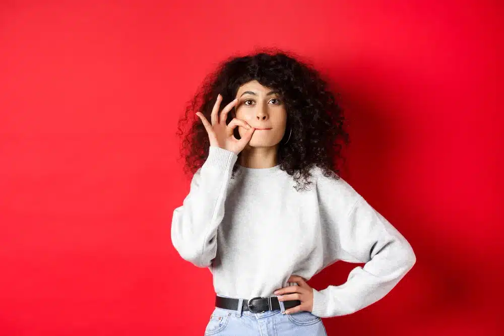 Estratégias para evitar deslizes verbais: o autocontrole no momento de falar Mulher com expressão pensativa, tocando o rosto e fazendo um gesto de 'silêncio', simbolizando reflexão e autocontrole verbal.