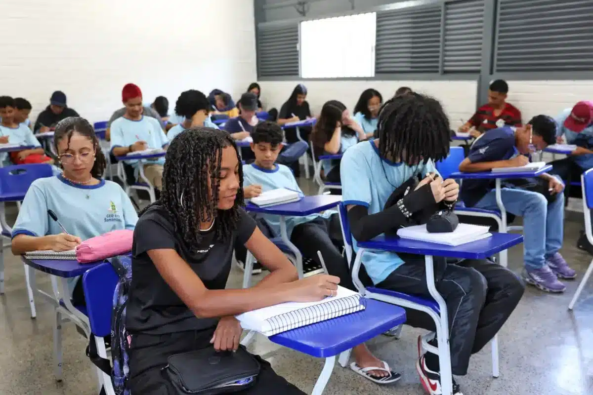 Pé-de-Meia Estudantes em sala de aula durante uma atividade escolar, todos com camisetas azuis, escrevendo em cadernos.