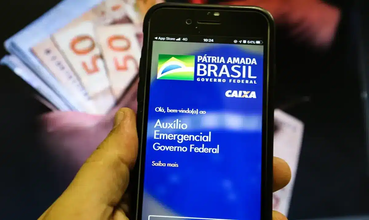 A lista de devolução do auxílio emergencial foi divulgada Celular exibindo a tela do Auxílio Emergencial, com cédulas de 50 reais ao fundo