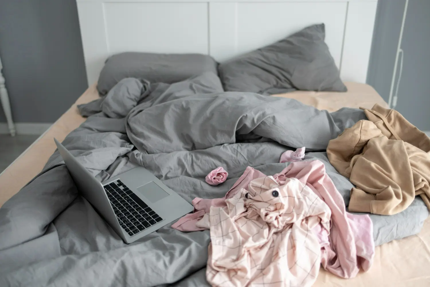 Cama desarrumada com roupas espalhadas e um laptop sobre a cama.