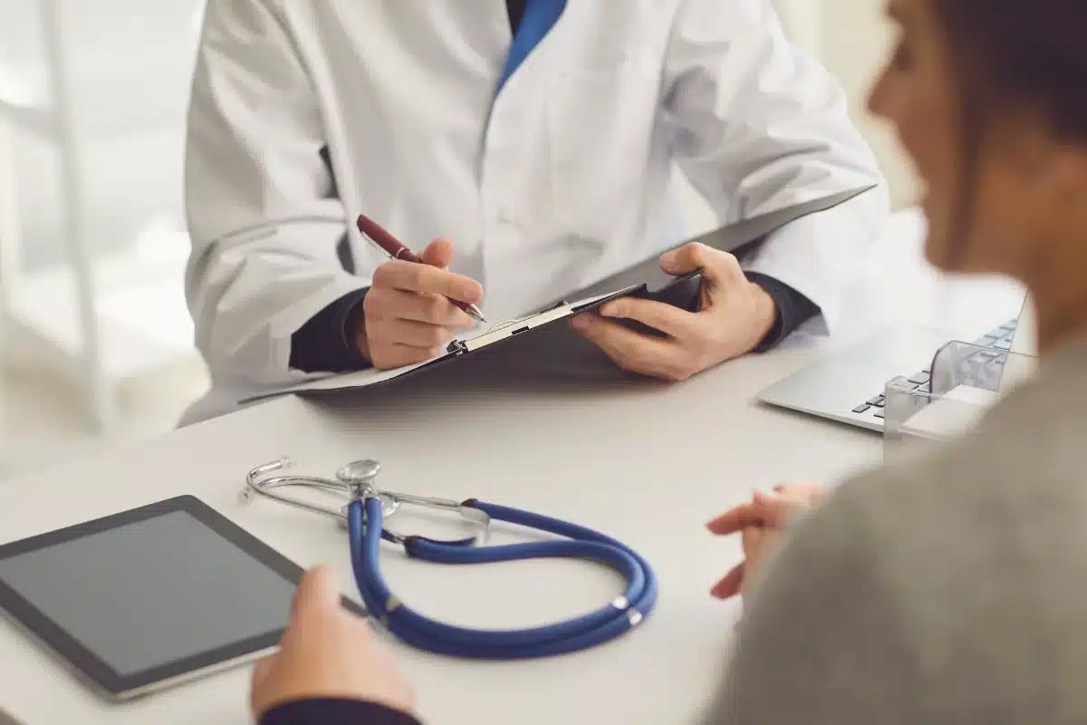Profissional da saúde Médico preenchendo um prontuário enquanto um paciente observa durante uma consulta.