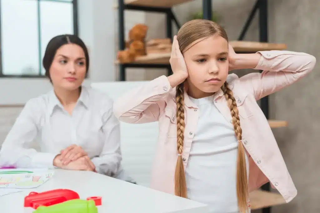 Como a escola pode identificar crianças com síndrome do imperador? Criança cobrindo os ouvidos enquanto uma professora observa, representando desafios na escola com a síndrome do imperador
