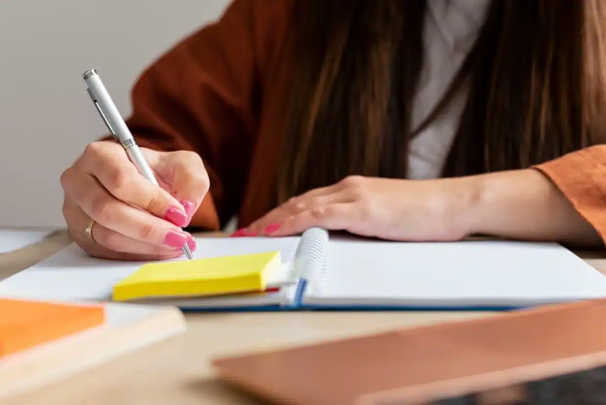 Pessoa anotando em caderno, refletindo sobre a importância do uso correto das expressões. Pessoa estudando, escrevendo em um caderno com post-its ao lado.