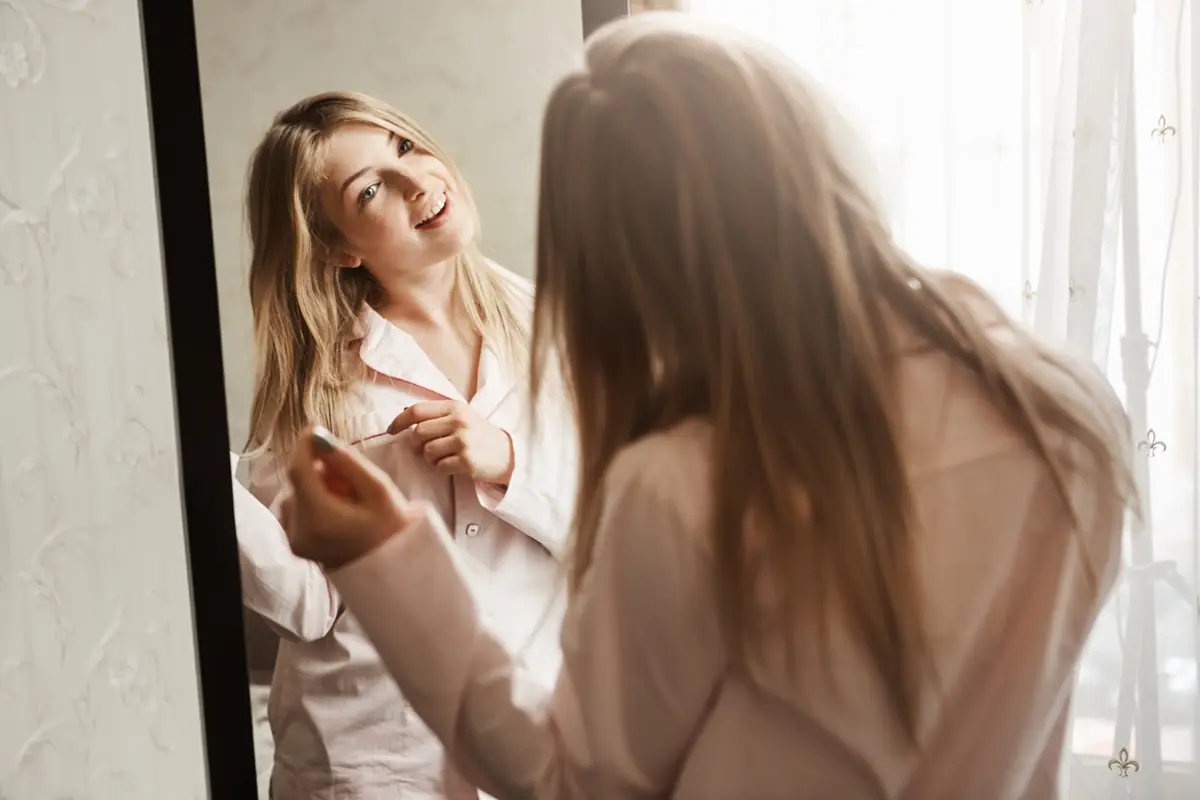 Mulher sorrindo e olhando para o espelho, refletindo autoestima e amor próprio.