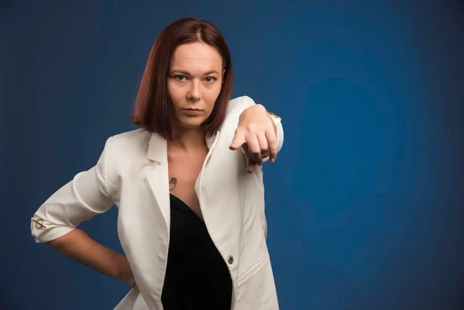 Mulher de expressão séria apontando diretamente para a câmera, vestindo blazer branco, em um fundo azul.