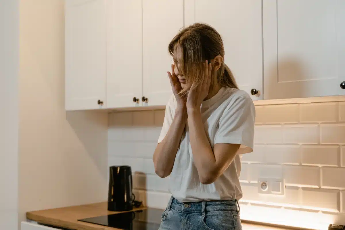 Mulher na cozinha com expressão de frustração, cobrindo o rosto com as mãos em um momento de estresse emocional.