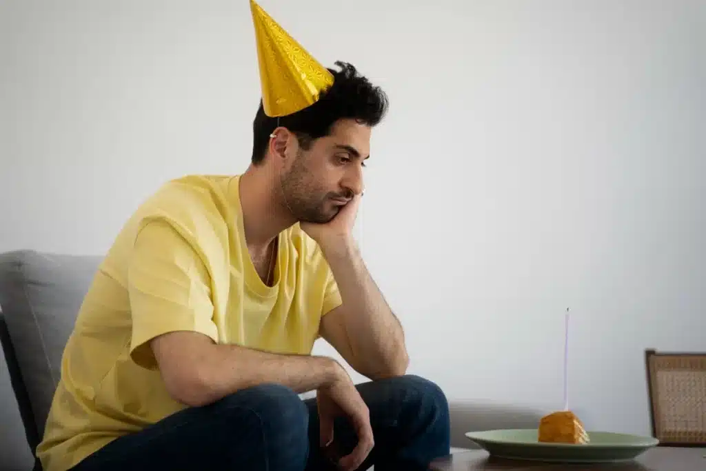 A autoestima tem grande impacto na comemoração do aniversário Homem com chapéu de festa, sentado sozinho, olhando para um bolo de aniversário, representando alguém que não gosta de comemorar seu aniversário.